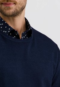 Marineblauer Strickpullover mit Rundhalsausschnitt, über einem dunkel gemusterten Hemd mit Kragen getragen, das weiße Blütenmuster auf einem marineblauen Hintergrund zeigt.