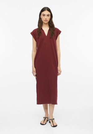 Femme debout portant une robe midi bordeaux sans manches avec un col en V et des sandales plates noires sur fond blanc.