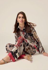 Satin-Outfit mit einer Bluse im floralem Muster in Schwarz, Rot und Beigetönen, kombiniert mit passenden Hosen und beigen flachen Schuhen.