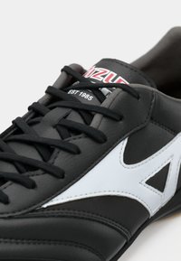 Pantofii athletic negri cu accente albe, superior din piele, șireturi negre plate și logo pe limbă. Caracterizează o textură netedă și detalii cusute.