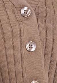 Beige geribbelde gebreide cardigan met metalen knopen met vier gaatjes en een gladde textuur. Close-up die de details van de knopen en de stof benadrukt.