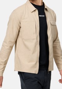 Homme portant une veste zippée beige sur un t-shirt noir avec texte blanc, pantalon foncé, debout les bras légèrement écartés sur fond uni.