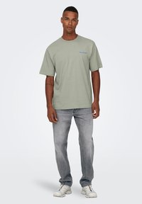 Camiseta de algodón verde clara con mangas cortas, cuello redondo y detalle gráfico azul en el pecho. Combinada con unos jeans grises desgastados y zapatillas blancas.