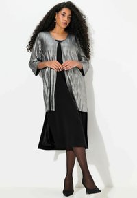Zwarte jurk met V-hals, kniehoogte, gelaagd onder een metallic zilveren cardigan met verticale strepen en wijde mouwen. Zwarte hakken.