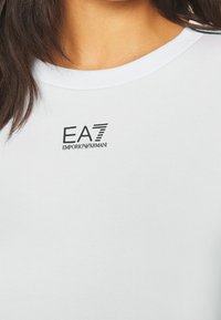 Primo piano di una persona che indossa una camicia bianca con il logo nero EA7 Emporio Armani stampato vicino al collo.