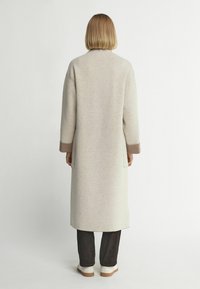 Beige kappa med långa ärmar, rak skärning och sidoklipp. Bruna ärmslut och subtil textur. Bärs över mörka byxor och vita skor.