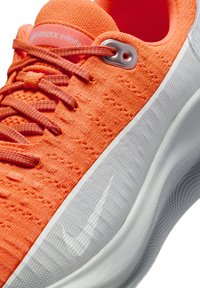 Scarpa da allenamento arancione brillante con tomaia in tessuto texturizzato, lacci a contrasto, occhielli argentati e intersuola bianca con logo Swoosh grigio.