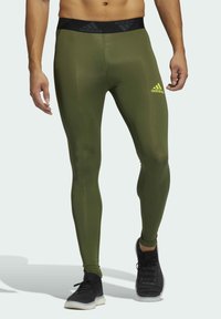 Oliwkowe legginsy o dopasowanym kroju, wykonane z elastycznego materiału, z czarnym paskiem w talii z logotypem "adidas" oraz małym żółtym logo.