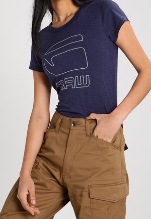 Kvinde iført en mørkeblå t-shirt med grafik, stoppet ind i brune cargobukser med høj talje, står med hånd i lommen mod en ensfarvet baggrund.