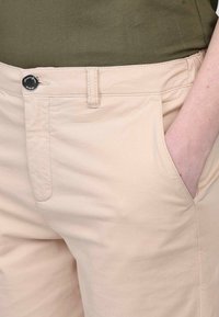 Pantalon beige en mélange de coton avec poches avant, présentant une fermeture à bouton et une texture lisse, partiellement visible au niveau de la taille avec un haut vert.