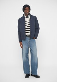 Ναυτικό καπιτονέ μπουφάν πάνω από λευκή και ναυτική ριγέ μπλούζα, με φαρδιά ανοιχτό μπλε τζιν και μαύρα παπούτσια, συμπληρωμένο με ένα μαύρο πλεγμένο σκουφάκι.