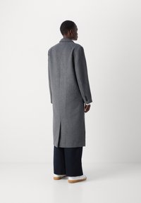 Palton lung din lână gri, cu un design ajustat, guler cu colțuri și o deschidere centrală în partea din spate. Purtați peste pantaloni largi bleumarin și adidași albi.