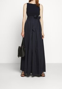 Kvinna iklädd en ärmlös marinblå maxi-klänning med en knuten midjebåge, hållande en svart bucket-väska och bärande svarta sandaler med öppen tå.