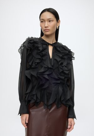 Femme portant un chemisier noir transparent à volants avec manches longues et une jupe en cuir marron, debout devant un fond clair uni.