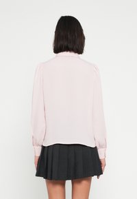 Ανοιχτό ροζ μπλούζα με μακριά μανίκια και γιακά, με λεπτομέρειες ραμμένες στους ώμους και στρογγυλεμένη τελείωση. Συνδυασμένη με σκούρα πλισέ φούστα.