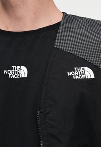 Κοντινό πλάνο ενός ατόμου που φοράει μαύρο μπλουζάκι και μπουφάν, και τα δύο με λευκά λογότυπα «The North Face» στο στήθος.