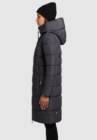 Dlhá vatovaná bunda tmavosivej farby s vysokým golierom a kapucňou. Obsahuje prešívané časti, vrecká so zipsom a trojuholníkový logový detail.