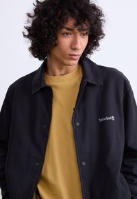 Black, lightweight jacket with button closures and Timberland logo, layered over a mustard yellow shirt, made from soft, textured fabric.