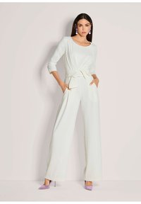Witte jumpsuit met driekwartmouwen, een ceintuur om de taille en een wijde pijp. In combinatie met lavendel hoge hakken en statement oorbellen.