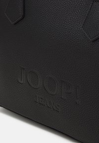 Črna teksturirana usnjena torba z izbočenim logotipom "JOOP! JEANS" in značilnimi heksagonalnimi kovinskimi detajli. Gladka površina.