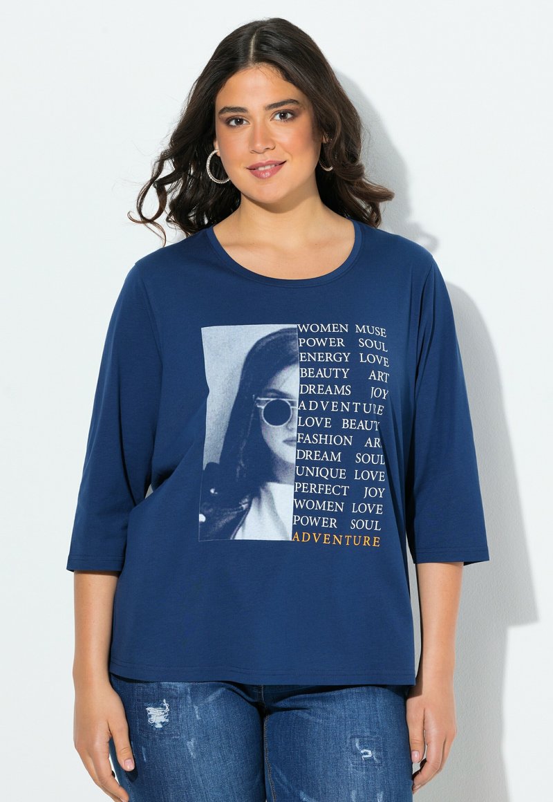 Námořnická modrá tričko s 3/4 rukávy, které obsahuje grafiku s bílým a oranžovým textem, znázorňující ženskou tvář ve slunečních brýlích. Měkká látka.