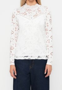 Blusa bianca in pizzo con maniche lunghe, motivi floreali, colletto alto e dettagli a bordo scallop, abbinata a pantaloni a gamba larga blu scuro.