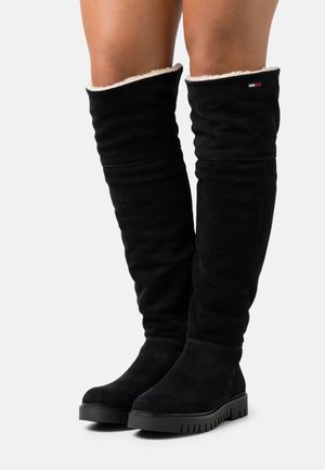 Black knee-high suede boots with white fleece lining worn on legs, featuring thick rubber soles and a small logo near the top edge.