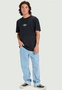 T-shirt preta de algodão com um logo geométrico colorido, combinada com jeans de denim azul claro e sapatilhas de lona pretas com solas brancas.