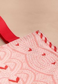 Punane kootud lõuendist kandekott, millel on roosa mustriga kujundus põimunud südametega, neid täiendavad tugevad punased sangad ja sile tekstuur.