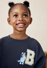 Dunkelblaues Sweatshirt mit einem Cartoon-Hundgrafik und einem flauschigen Buchstaben "B"-Patch. Weicher Stoff mit geripptem Rundhalsausschnitt und lockerer Passform.