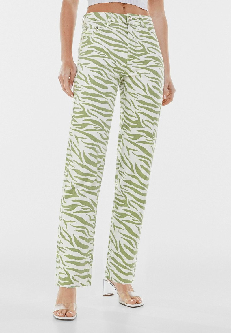 Jeans à jambes larges imprimé zèbre vert et blanc, taille haute, avec fermeture à boutons et zip, ainsi que des poches latérales. Sandales à talons transparents.