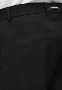 Μαύρο παντελόνι με ραφές, με τσέπη με λωρίδα στο πίσω μέρος και ορατή μαύρη ετικέτα στη μέση, σε άτομο που φοράει λευκό υφασμάτινο τοπ.