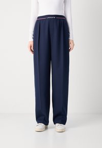 Pantaloni a gamba larga blu navy con vita alta caratterizzati da un logo Lacoste e una striscia accentuata. Tessuto liscio, pieghe frontali e vestibilità rilassata.