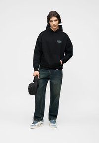 Mlada oseba, ki nosi črno kapuco, temne kavbojke in bele superge, drži majhno črno torbico in ima eno roko v žepu.