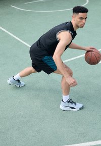 Black sleeveless jersey with white trim, teal shorts, gray basketball shoes, dribbling a basketball on a green outdoor court.