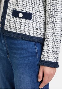 Weißes und marineblaues Tweed-Jackett mit Fransenverzierungen, Knopfschluss, strukturiertem Stoff und einer kontrastierenden marineblauen Taschenakzent. Getragen mit blauen Jeans.