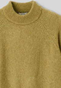 Pull en maille dans un vert olive clair avec un col roulé côtelé. Texture douce, manches raglan et une étiquette visible à l'intérieur du col.