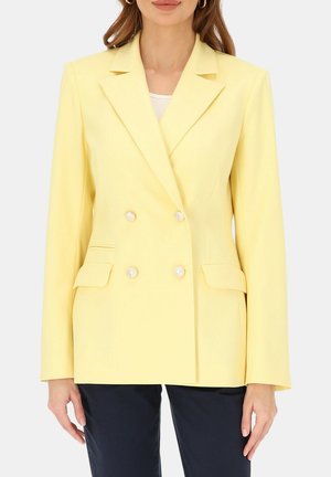 Kobieta w bladożółtej dwurzędowej marynarce z czterema ozdobnymi guzikami i klapowanymi kieszeniami, w połączeniu z ciemnymi spodniami.