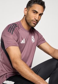 Μωβ αθλητική μπλούζα με μαύρες ρίγες, που φέρει λευκό λογότυπο στο στήθος. Συνδυάζεται με μαύρο παντελόνι, αναδεικνύοντας ένα αθλητικό σχέδιο και εφαρμογή.