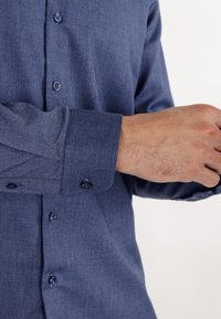 Marineblå skjorte med knapper, teksturert finish, lange ermer og knappet mansjett. Har matchende knapper langs fronten.