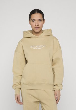 Νέος/Νέα φοράει μπεζ φούτερ Rocawear με κουκούλα και ταιριαστό παντελόνι, στέκεται μπροστά σε απλό λευκό φόντο.
