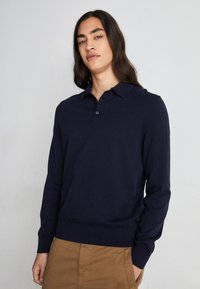 Ναυτική πόλο μπλούζα με μακριά μανίκια, κουμπωτό γιακά, ραμμένα μανίκια και στρίφωμα, φτιαγμένη από μαλακό ύφασμα, συνδυασμένη με ανοιχτό καφέ παντελόνι.