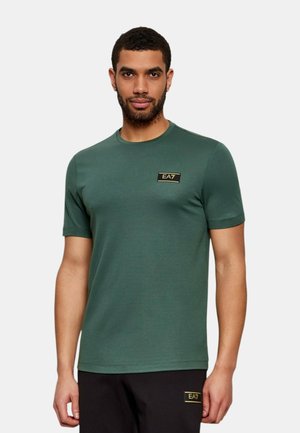 Hombre que lleva una camiseta verde de manga corta con el logo EA7 en el pecho y pantalones negros con el logo EA7 en el muslo, de pie frente a un fondo liso.