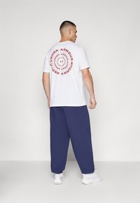 White cotton t-shirt with red circular "Under Armour" graphic on the back; paired with loose, dark blue pants and white sneakers.