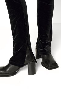 Pantalon en velours noir avec fentes latérales porté avec des bottines noires à talon carré sur fond blanc.