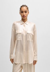 Helles Beige, durchscheinendes Langarmshirt mit einem Hauch von Schimmer, zwei Brusttaschen und einem klassischen Kragen. Aufweist eine entspannte Passform und eine strukturierte Oberfläche.