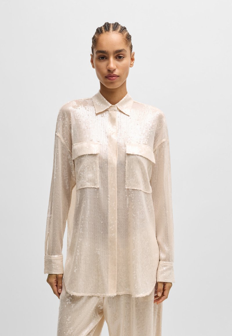 Helles Beige, durchscheinendes Langarmshirt mit einem Hauch von Schimmer, zwei Brusttaschen und einem klassischen Kragen. Aufweist eine entspannte Passform und eine strukturierte Oberfläche.