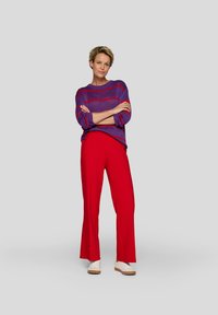 Purple and red striped knit sweater, red wide-leg trousers, and white sneakers. Model stands with arms crossed against a gray background.
