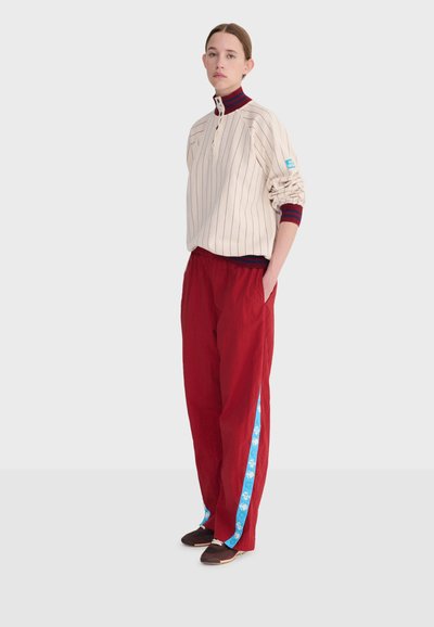 Jeune personne debout, portant une chemise à manches longues crème rayée avec un col et des poignets rouges et bleus, un pantalon rouge avec des bandes latérales bleues, des chaussures marron.