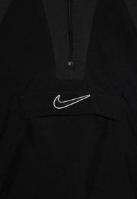 Crna zip-up majica s teksturiranim materijalom na kojem se nalazi bijeli Nike logo na prednjem džepu, naglašavajući pojednostavljen dizajn.